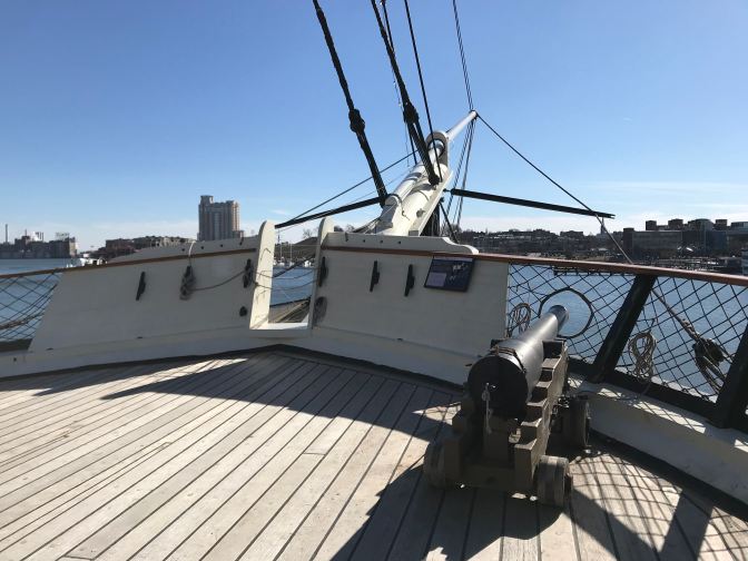 Bow of ship, with bowsprit extending outward, and a cannon on deck in the foreground.