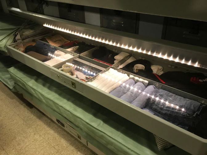 Storage locker beneath a bunk bed, showing towels, shirts, pants, and various personal articles.