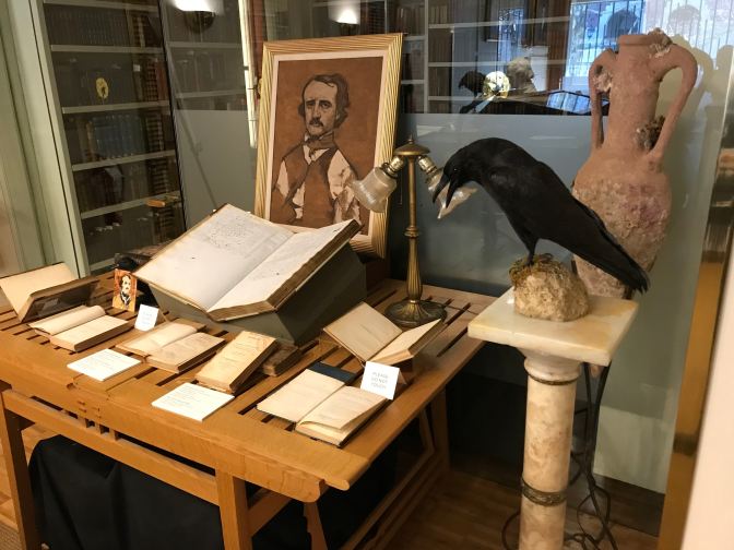 Table with books and a painting of Edgar Allan Poe. A raven doll is on a perch in the foreground.