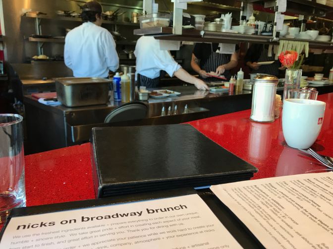 Menu on red countertop in foreground. Chefs work in open kitchen of restaurant in background.