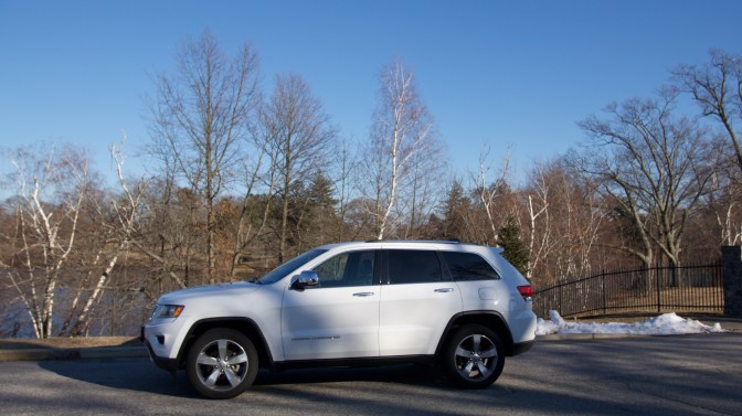2014 white Jeep Grand Cherokee, parked in front of trees near a fence. Some snow is on the ground.