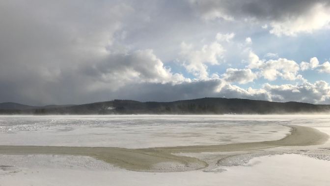 View of frozen lake, with mountains in the background and a cloudy sky above.