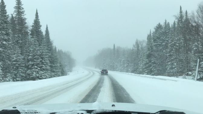 View of mountain road during snowstorm. Trees line the road on both sides, and a car is in front.