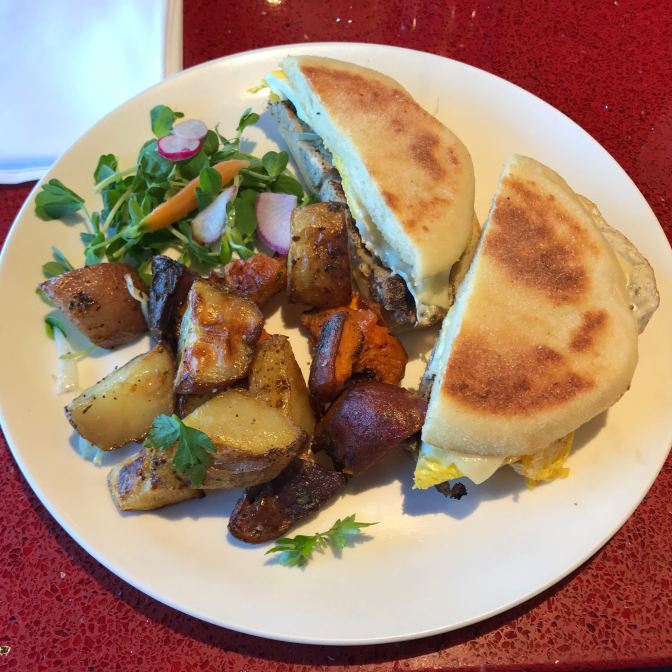Chicken sausage and egg on an english muffin, with roasted potatoes and greens, on a white plate.