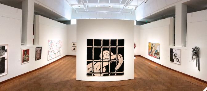 Panorama of ground level gallery, with a large drawing of an octopus tentacle behind a prison cell in the middle of the room.
