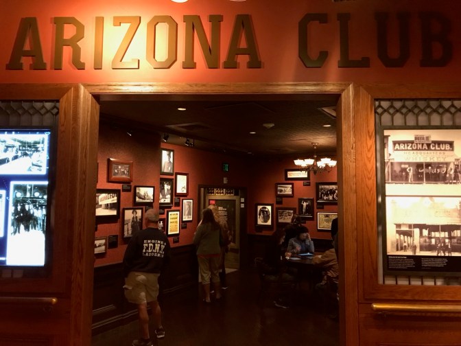 Exterior of hotel room with the words ARIZONA CLUB across the top of the doorway. The view through the open doorway shows a replica casino with pictures hanging on the wall.