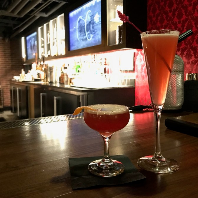 Long stem glass with cocktail, and shorter rounded glass with cocktail on bar.