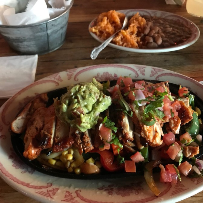 Plate with chicken, pico de gallo, guacamole, and peppers. In the background is another plate with rice and beans.