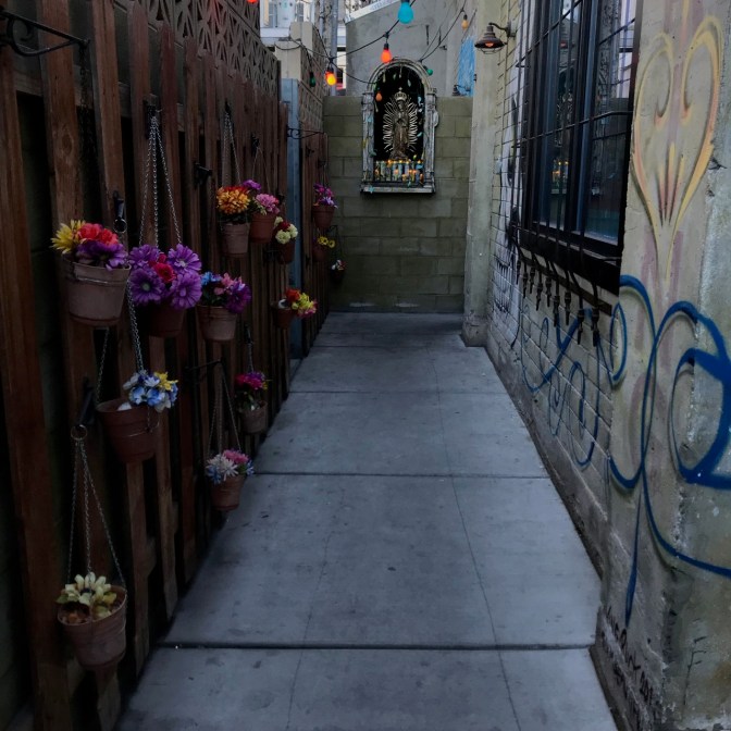 Exterior entrance of La Comida. A small shrine to Mary is located in a grotto in a wall, and plants hang from a wooden fence.