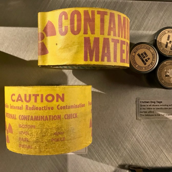 Caution tape and civilian dog tags. The caution tape says CONTAMINATED MATERIAL CAUTION INTERNAL RADIOACTIVE CONTAMINATION.