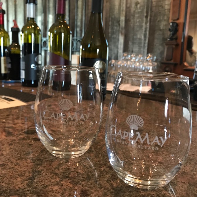 Two glasses with CAPE MAY WINERY & VINEYARD engraved on the glass. Wine bottles are in the background on the bar top.