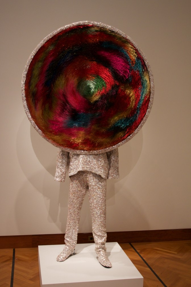 Statue of man with large circle where head should be. Garment is made out of buttons.