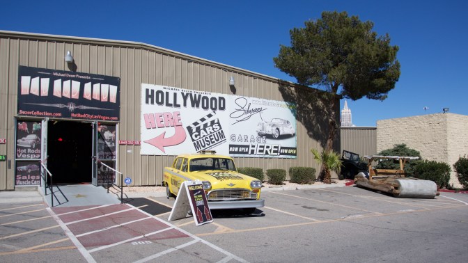 Exterior of Hollywood Cars Museum, with a yellow cab and the Flinstone's car parked in front of the building. A large sign says HOLLYWOOD CARS MUSEUM HERE.