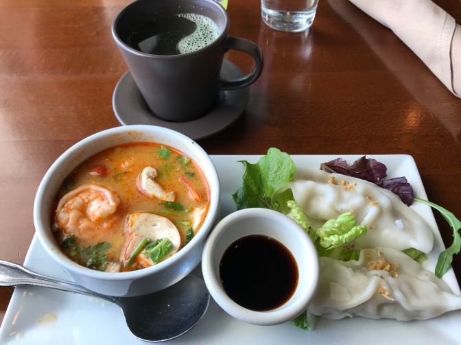 White rectangular plate with bowl of tom yum soup, a smaller bowl of soy sauce, and two pork dumplings.