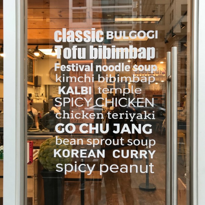 Glass door with words CLASSIC BULGOGI TOFU BIBIMBAP FESTIVAL NOODLE SOUOP KIMCHI BIBIMBAP KALBI TEMPLE SPICY CHICKEN CHICKEN TERIYAKI GO CHU JANG BEAN SPROUT SOUP KOREAN CURRY SPICY PEANUT