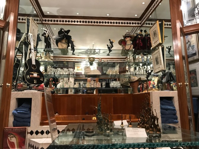 Elaborate bathtub, which chess set on glass table in front of tub. A guitar, statues, and glasses are on glass shelves behind the tub.