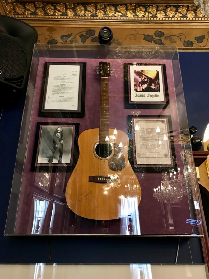 Glass case with Janis Joplin's acoustic guitar, a photo of Janis, her will, and her death certificate.