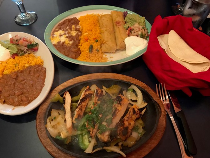 Tamales on plate with rice and refried beans. Fajita sizzling on skillet. Tortillas in basket with red cloth. Plate with lettuce, guacamole, refried beans, and rice, all on a table.