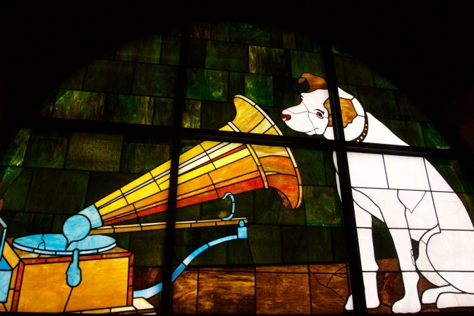 Stained glass window of white dog listening to record player.