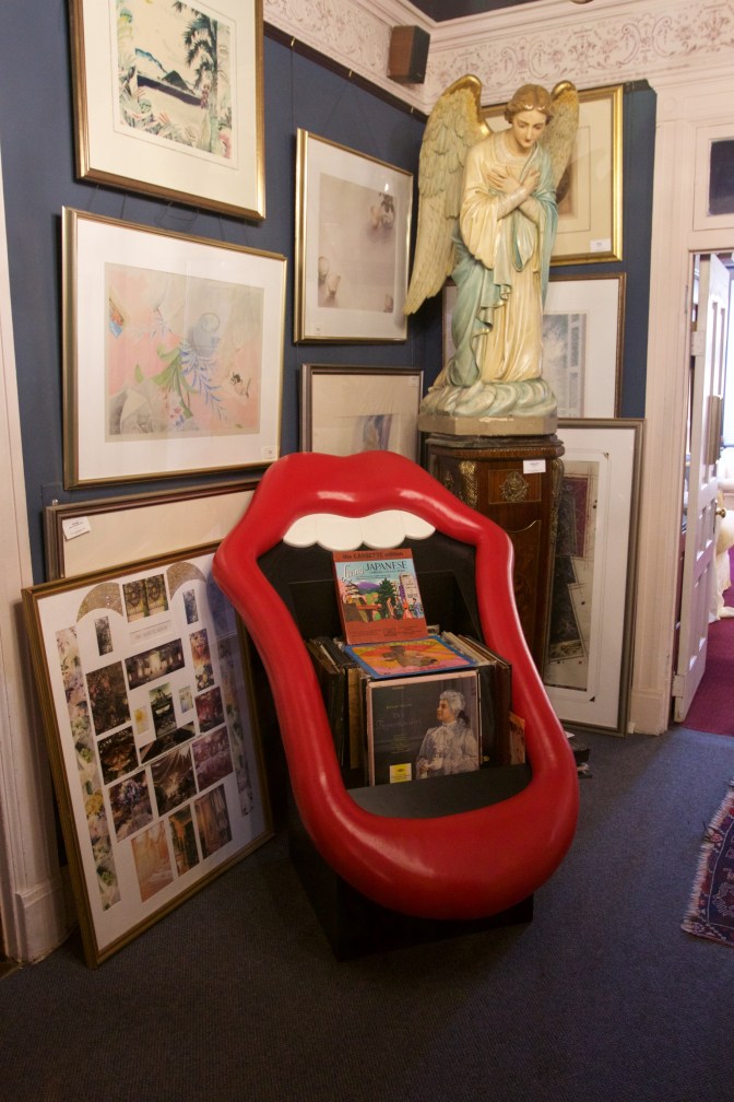 Red lips and mouth chair, angel statue, and artwork, in a corner of a room.