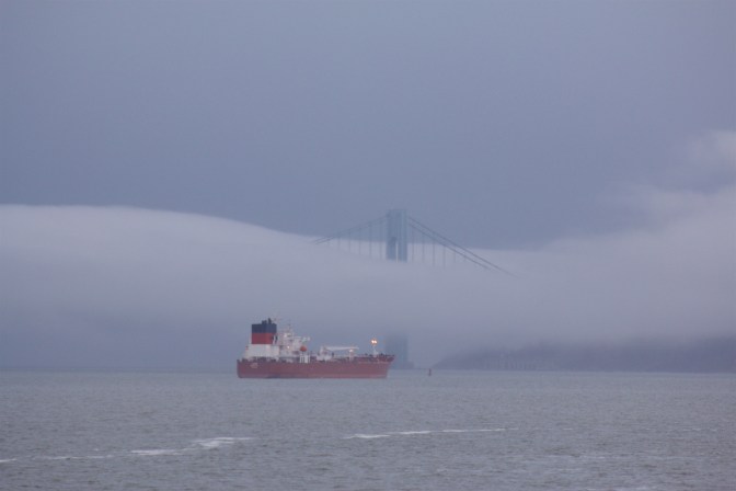 Fog obscures the Verrazano Narrows bridge, with a freighter in the foreground.