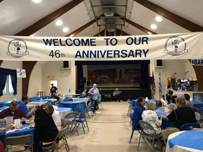 Church hall that has a sign that says WELCOME TO OUR 46th ANNIVERSARY. Musicians play at the front, while people sit at round tables.