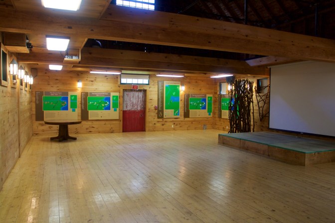Interior of barn, with a stage, a screen, and maps of the evolution of Camp Sagamore.