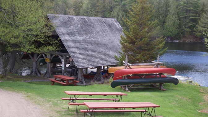 Boathouse, with canoes on a rack outside of it, and picnic tables in the foreground.