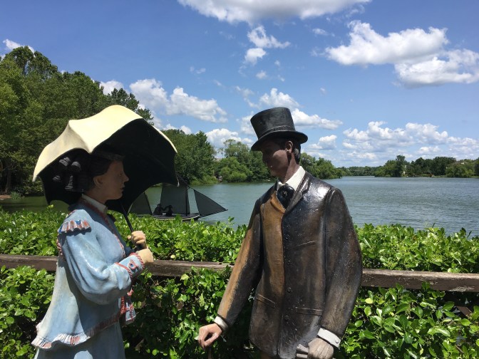 Man in suit and top had and woman in dress and parasol beside lake.