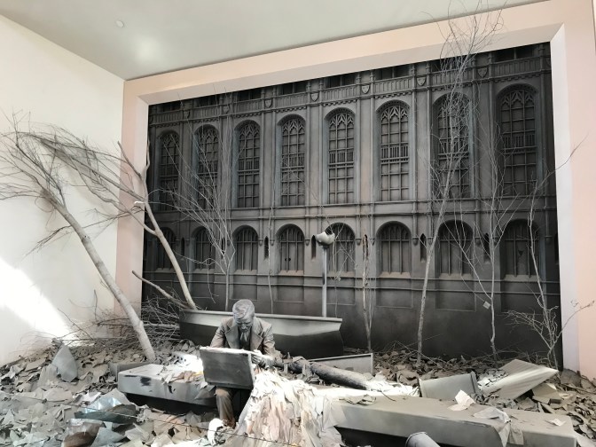 Sculpture of man looking in briefcase, with rubble and dead trees surrounding him. A building facade is in the background.