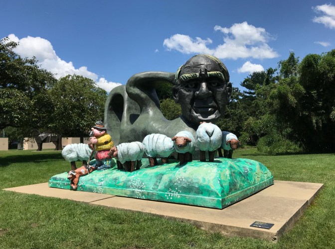 Sculpture of man's head surrounded by sheep and small shepherd.
