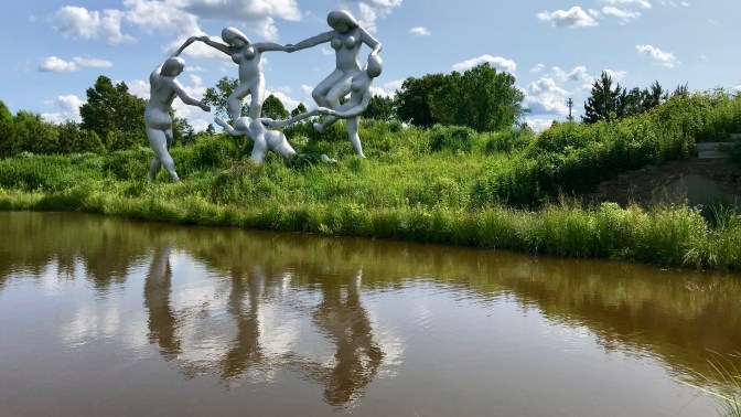 Five large statues of dancers by a lake.