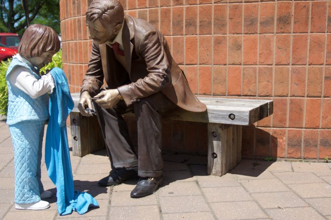 Sculpture of man and child. The man is seated on a bench, the child is holding a blanket, standing in front of the man.