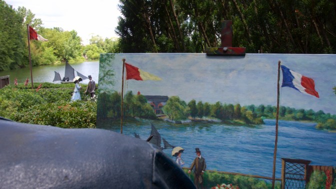 Easel with painting of lake, with lake scene in background.