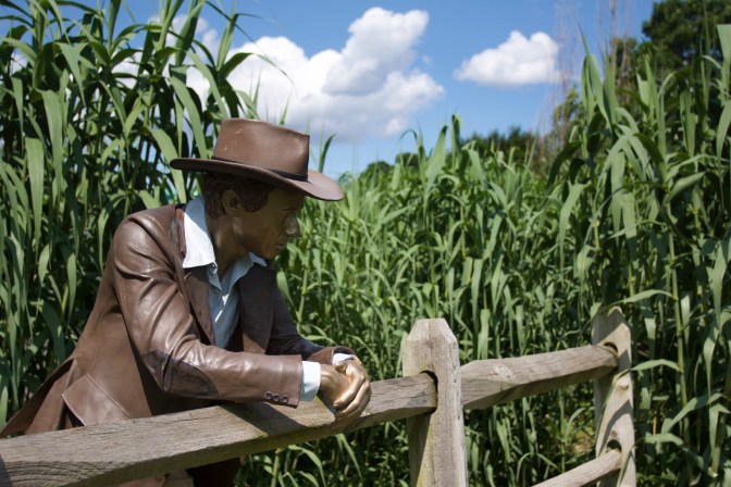 Sculpture of man standing at wooden fence, with corn behind him.