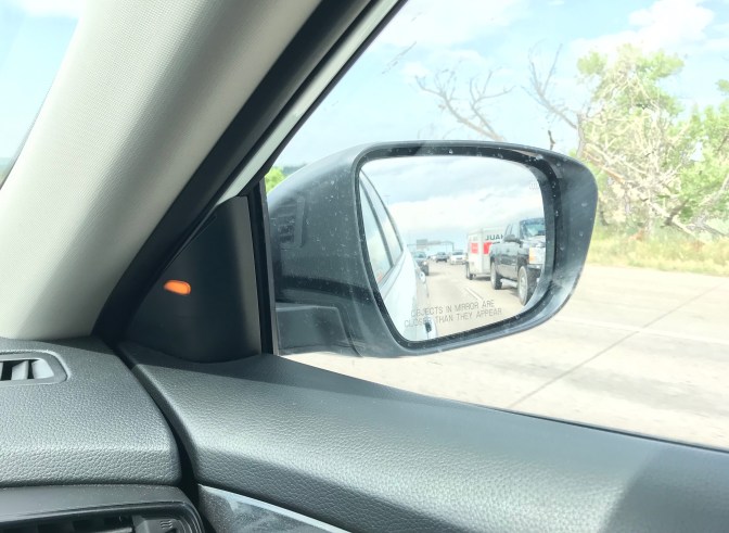 Rearview mirror of car, with yellow light flashing beside it. A truck is visible in the mirror.
