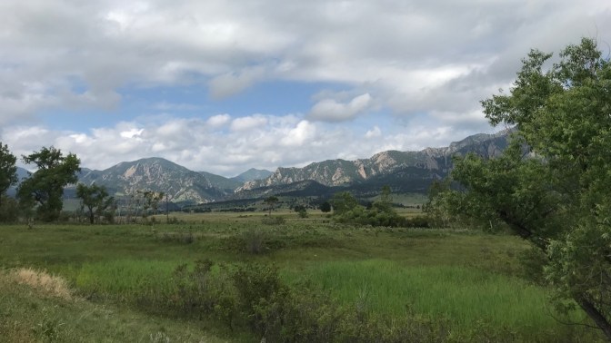 Trees and grassland in foreground, and foothills in background beneath a cloudy sky.
