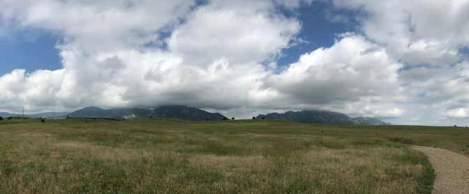Panorama of foothills of Rocky mountains, with clouds in the sky and a grassy prairie in the foreground.