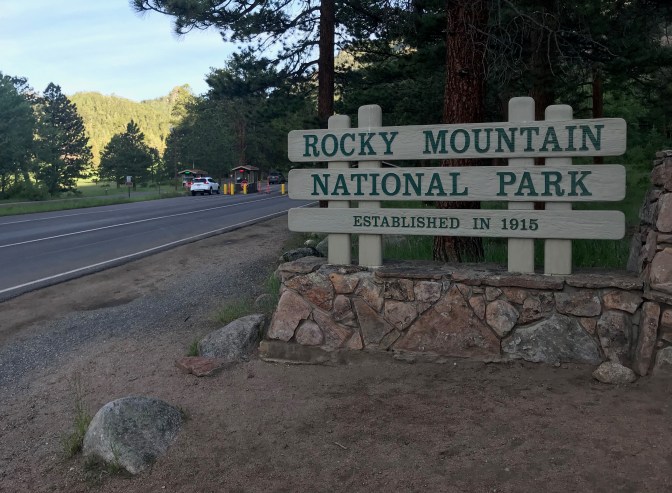 Entrance road to the park, with a large sign in foreground that says ROCKY MOUNTAIN NATIONAL PARK ESTABLISHED IN 1915.
