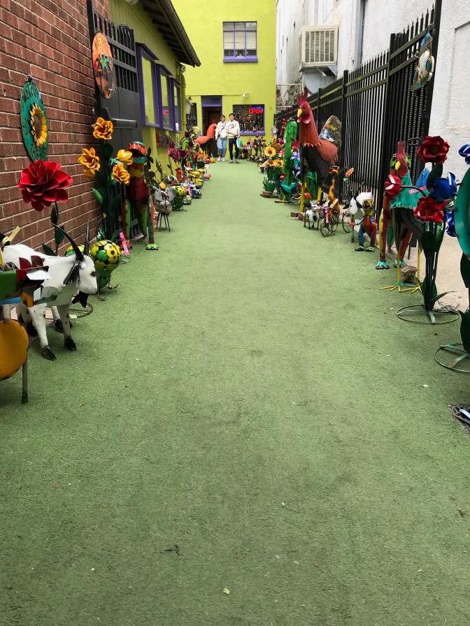 Small metal statues in alley with yellow building in background.