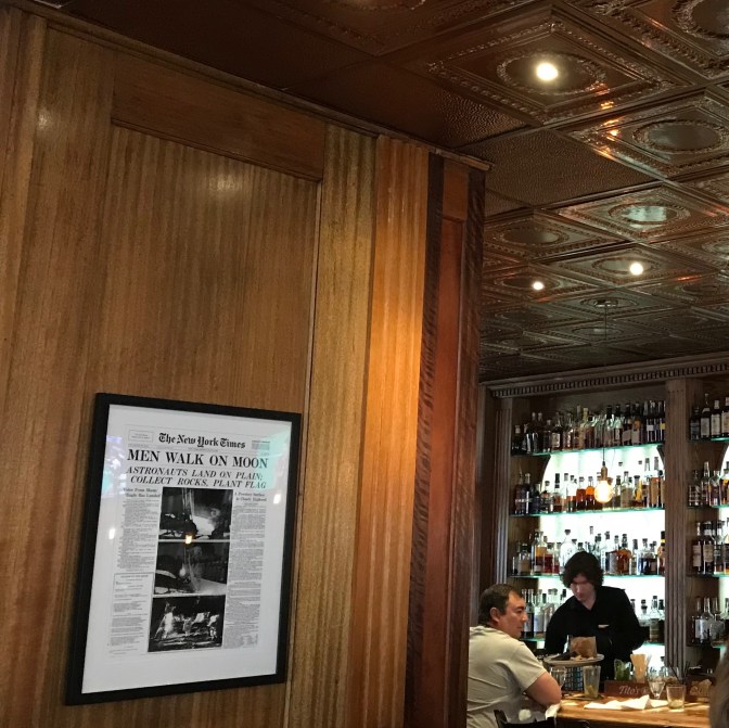 Replica of New York Times with front-page stating MEN WALK ON MOON, hanging on wall in frame. The restaurant bar is in background.