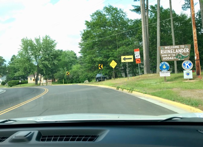 View of street from behind dashboard of car. Sign on right of road says WELCOME TO RHINELANDER HOME OF THE HODAG.