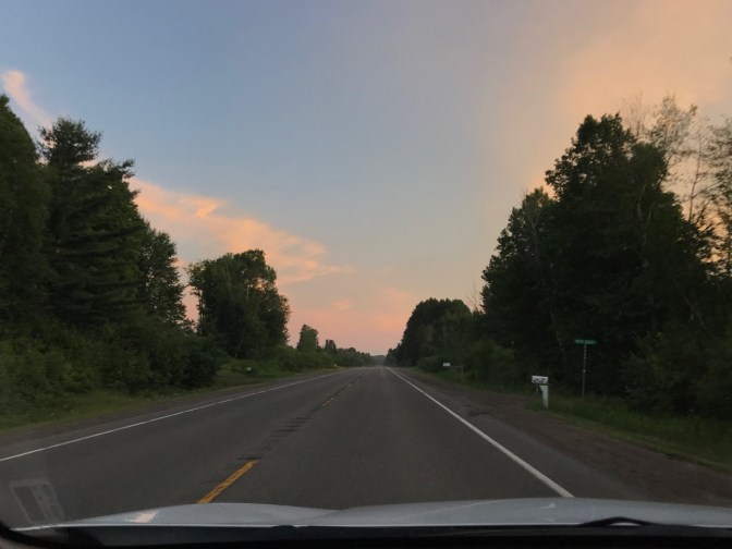 View of sky at dusk through car windshield. A tree-lined road is in the foreground.