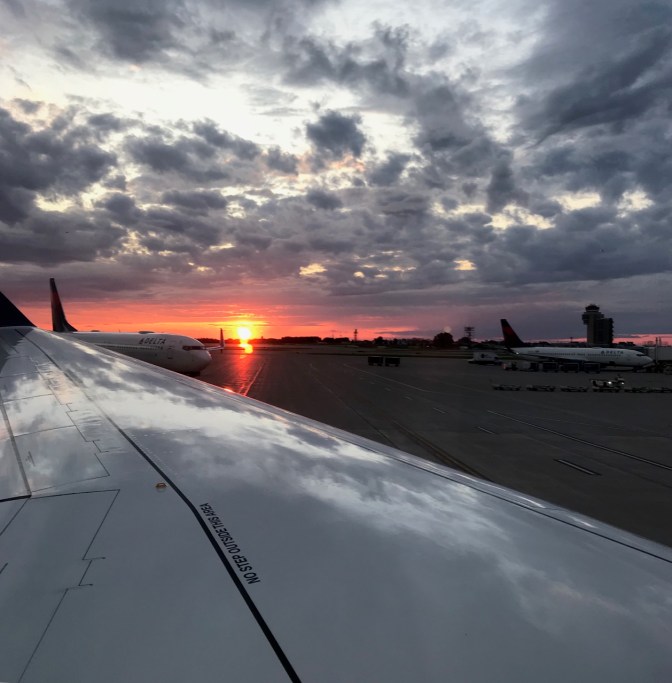 View of sunset over wingtip of airplane parked at airport. Other airplanes are in the background.