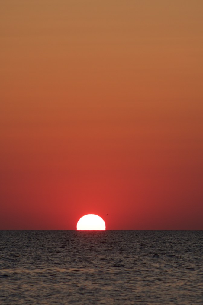 Setting sun with ocean in foreground and an orange sky.