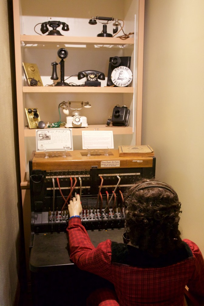Mannequin at telephone switchboard, with various telephones on shelves above.