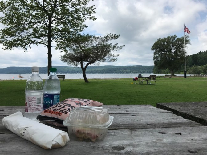 Two bottles of water, two sandwiches, and a bag of chips on a picnic table beside a lake.