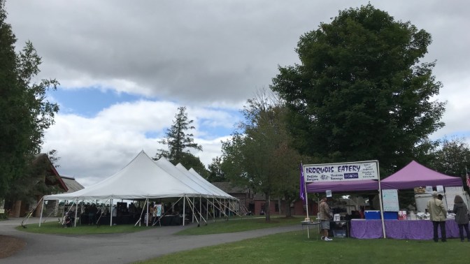 White tent with arts and crafts on left of field and a tent on the right selling food. A sign on the right tent says IROQUOIS EATERY.