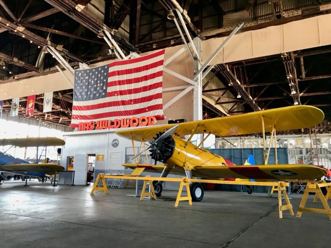 Interior of Naval Air Station hangar, with a yellow biplane in foreground and an American flag hanging from the ceiling.