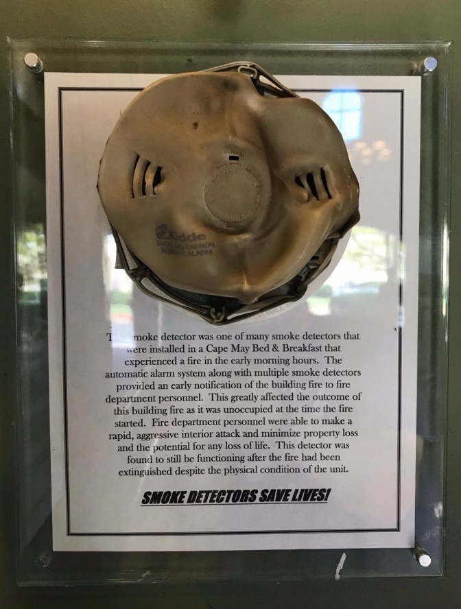 Melted smoke detector on plaque. Sign says THIS SMOKE DETECTOR WAS ONE OF MANY SMOKE DETECTORS THAT WERE INSTALLED IN A CAPE MAY BED AND BREAKFAST THAT EXPERIENCED A FIRE IN THE EARLY MORNING HOURS. THE AUTOMATIC ALARM SYSTEM ALONG WITH MULTIPLE SMOKE DETECTORS PROVIDED AN EARLY NOTIFICATION OF THE BUILDING FIRE TO FIRE DEPARTMENT PERSONNEL. THIS GREATLY AFFECTED THE OUTCOME OF THIS BUILDING FIRE AS IT WAS UNOCCUPIED AT THE TIME THE FIRE STARTED. FIRE DEPARTMENT PERSONNEL WERE ABLE TO MAKE A RAPID AGGRESSIVE INTERIOR ATTACK AND MINIMIZE PROPERLY LOSS AND THE POTENTIAL FOR ANY LOSS OF LIFE. SMOKE DETECTORS SAVE LIVES.